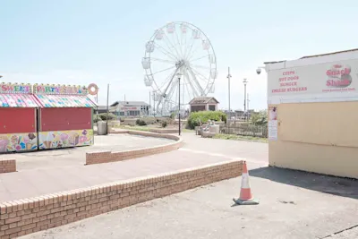 Large white Ferris wheel at a seaside amusement area with food stalls and brick walkways on a sunny day.