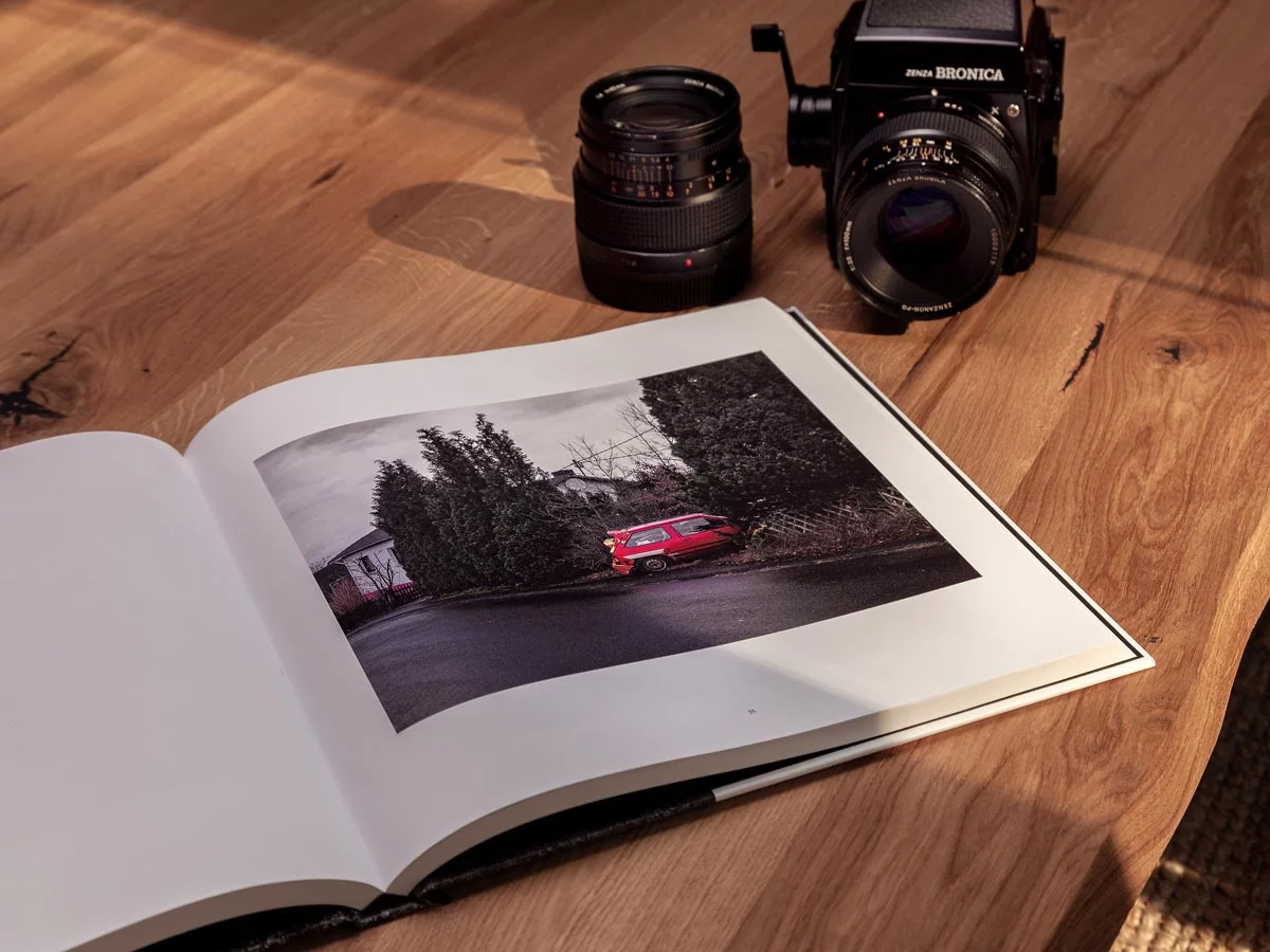 An illustrated book lies on a wooden table, open with a photo of a red car in front of bushes, next to it a camera and a lens.