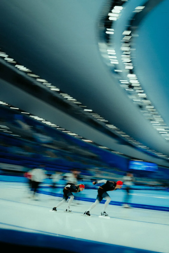 A dynamic shot of two speed skaters in a tucked position during a race. The image conveys high speed through intentional motion blur. In the background, the curved stands and bright stadium lighting of a modern indoor ice arena are visible.