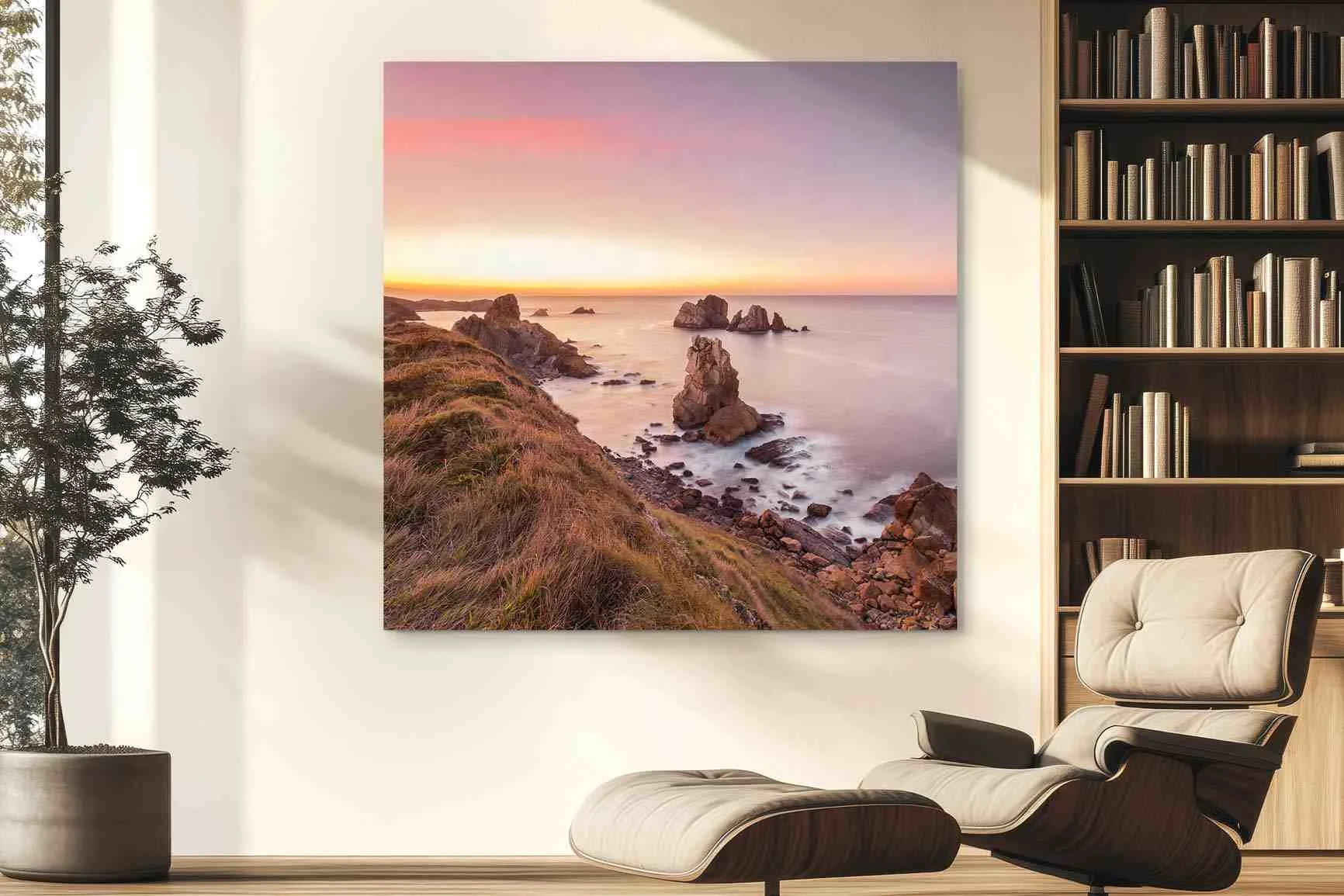Stampa di grande formato raffigurante un paesaggio costiero con scogli nel mare al tramonto su una parete chiara sopra una poltrona e una libreria.