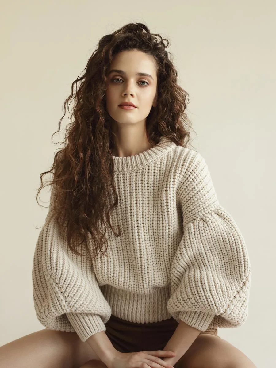 A woman with long curly hair wears a cream-colored, coarsely knitted sweater with wide sleeves. She is sitting in front of a beige background.