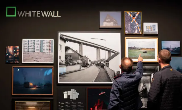 Two people stand in front of a black wall with a WhiteWall logo and look at a selection of framed photographs in various formats and styles.