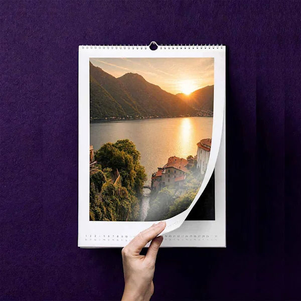 Una mano sfoglia un calendario fotografico con un’immagine paesaggistica di un lago e delle montagne al tramonto.