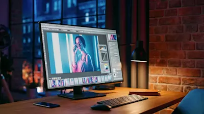 Monitor zeigt Bildbearbeitungssoftware mit Porträtfoto, daneben Tastatur, Lampe und Handy auf Schreibtisch.