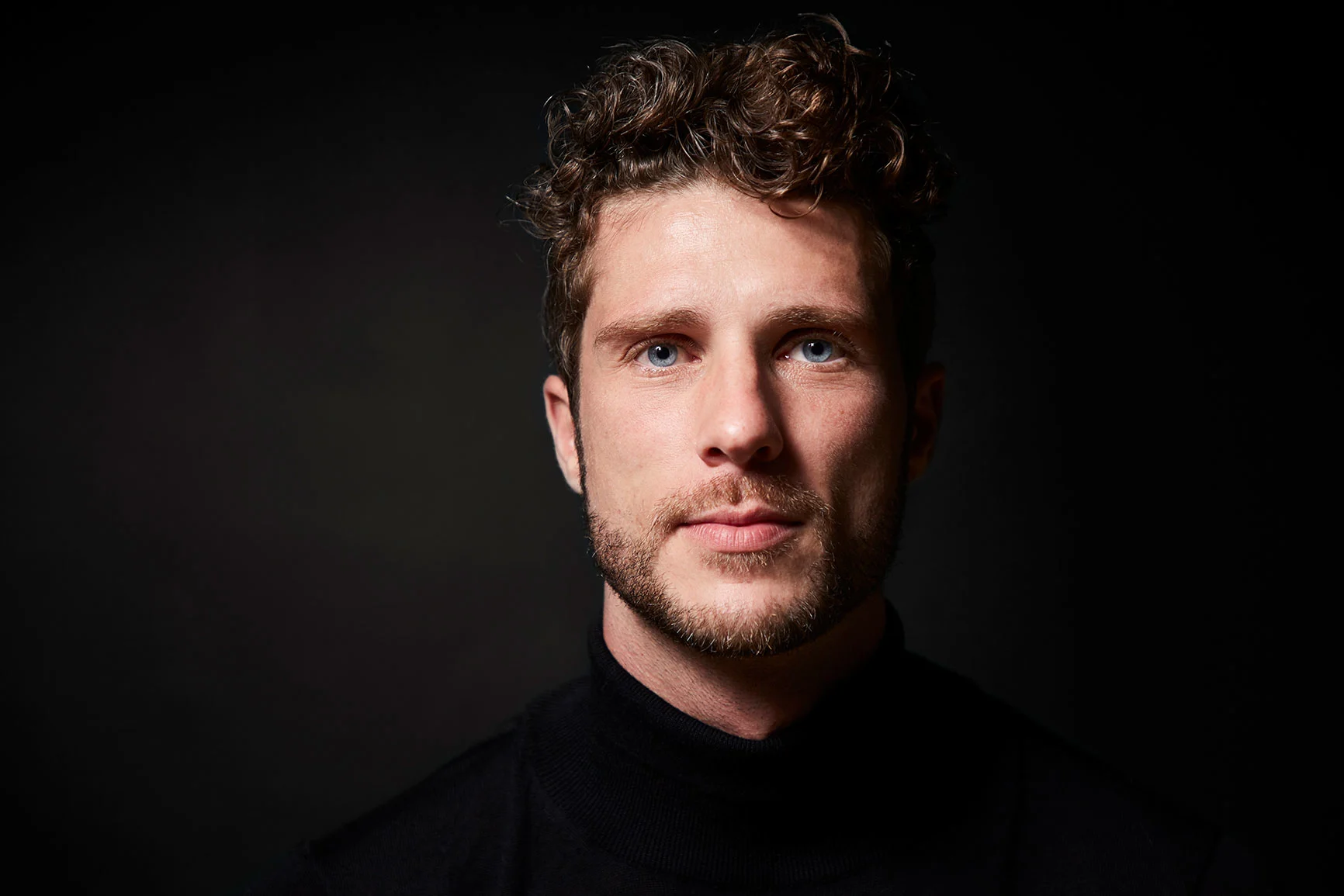 A man with curly hair and a black turtleneck sweater against a black background.