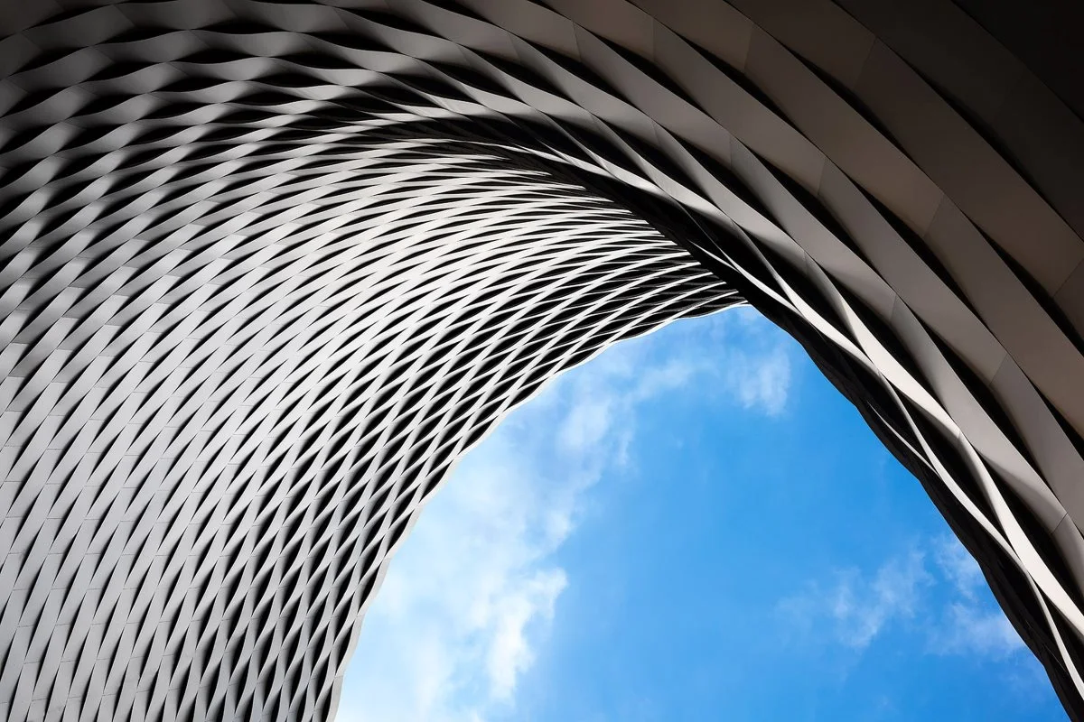 Moderne architektonische Detailaufnahme einer geschwungenen weißen Metallgitterstruktur vor strahlend blauem Himmel mit zarten Wolken