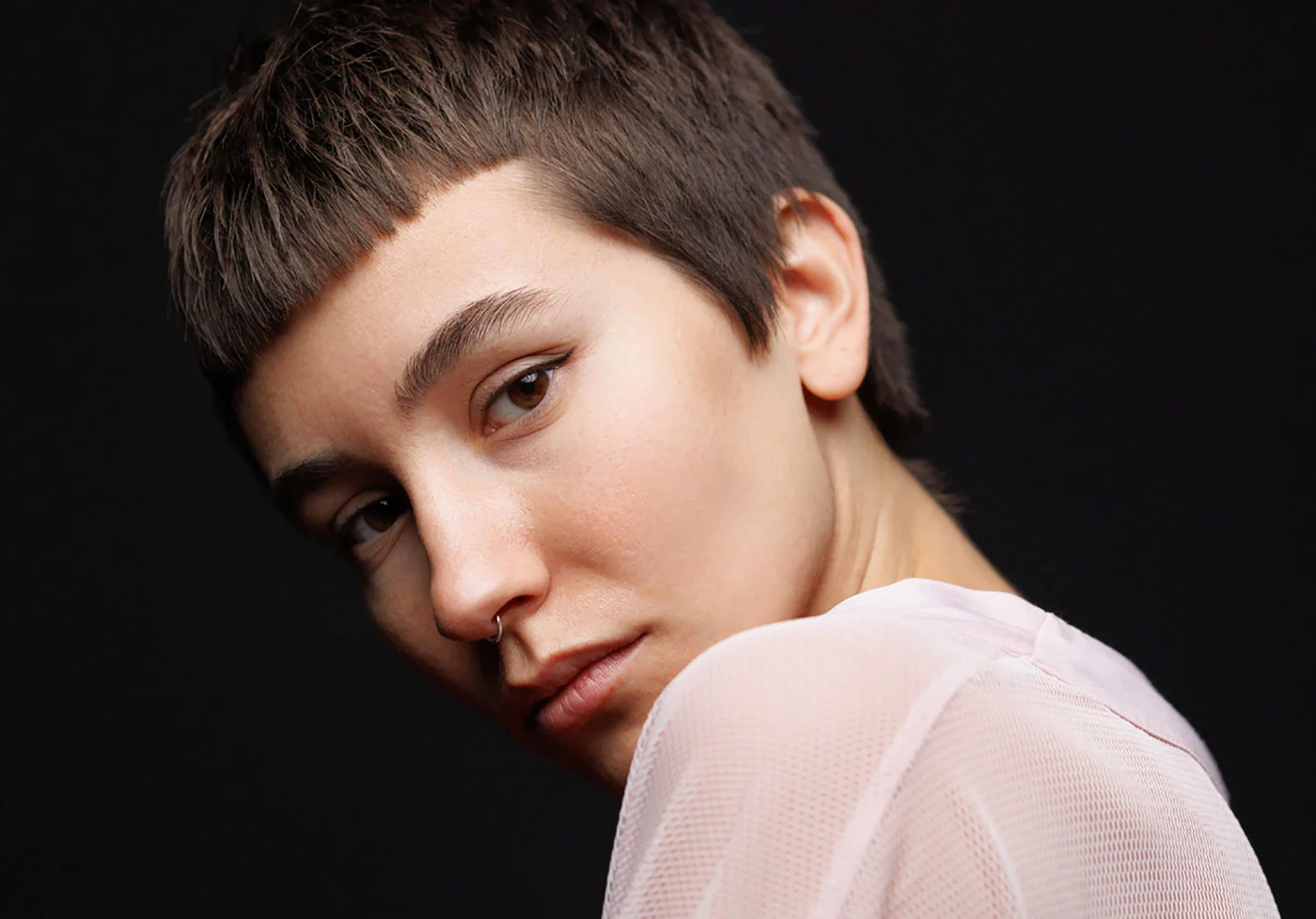Person with short brown hair wearing a white, transparent top against a black background.