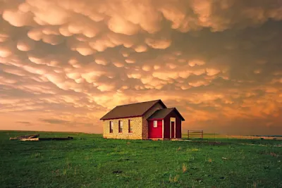 Ein kleines Bauernhaus aus Stein und rotem Holz steht auf einer grünen Prärie unter einem dramatischen orangefarbenen Himmel, der von Mammatuswolken erfüllt ist.