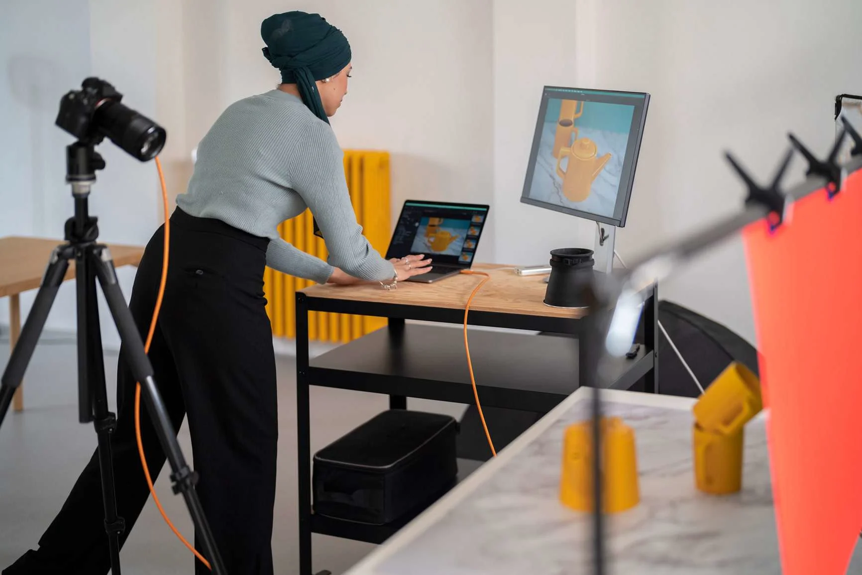 A photographer at a laptop uses a cable to control a camera on a tripod for a product shoot of yellow jugs in the studio.