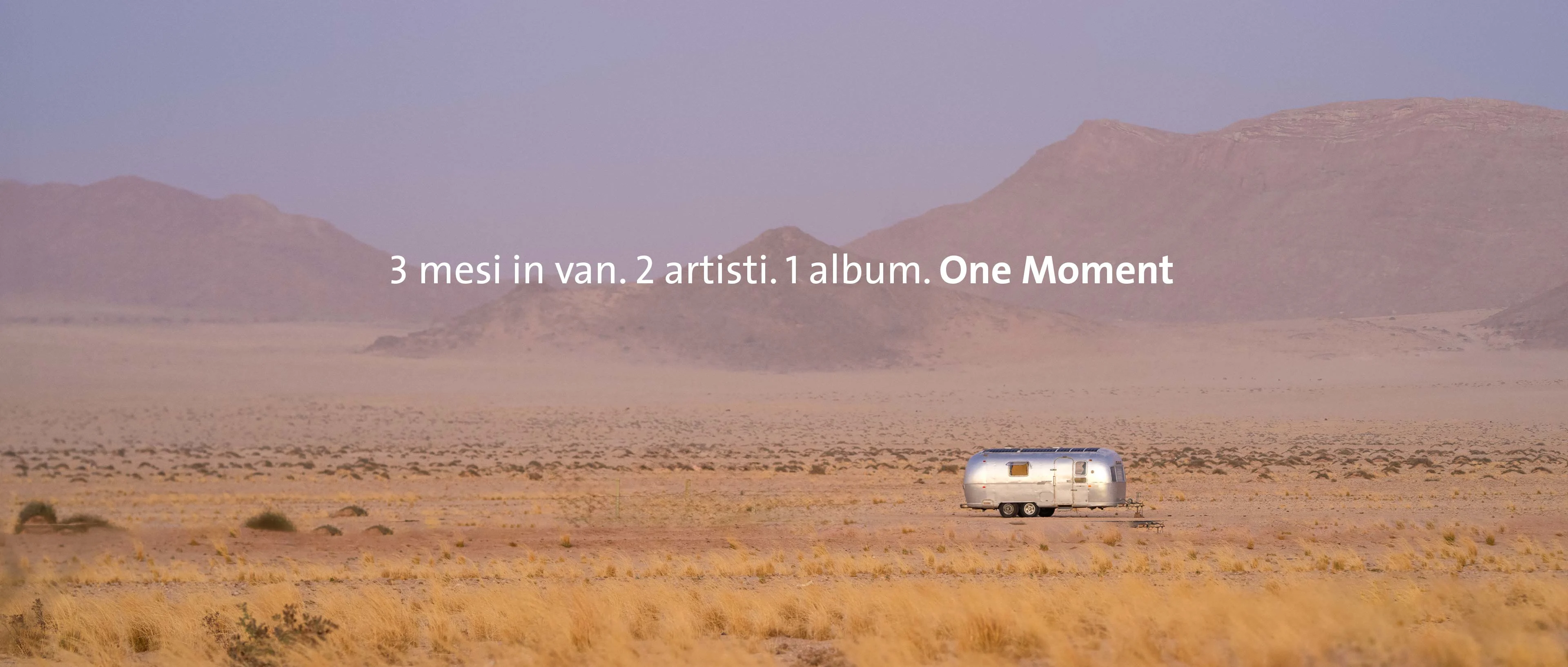 Un'ampia inquadratura panoramica del deserto namibiano in una luce viola diffusa. In mezzo alla pianura sabbiosa si trova un camper Airstream d'argento davanti a una dolce catena montuosa. Al centro dell'immagine campeggia il testo: "3 mesi in un furgone. 2 artisti. 1 album. One Moment".
