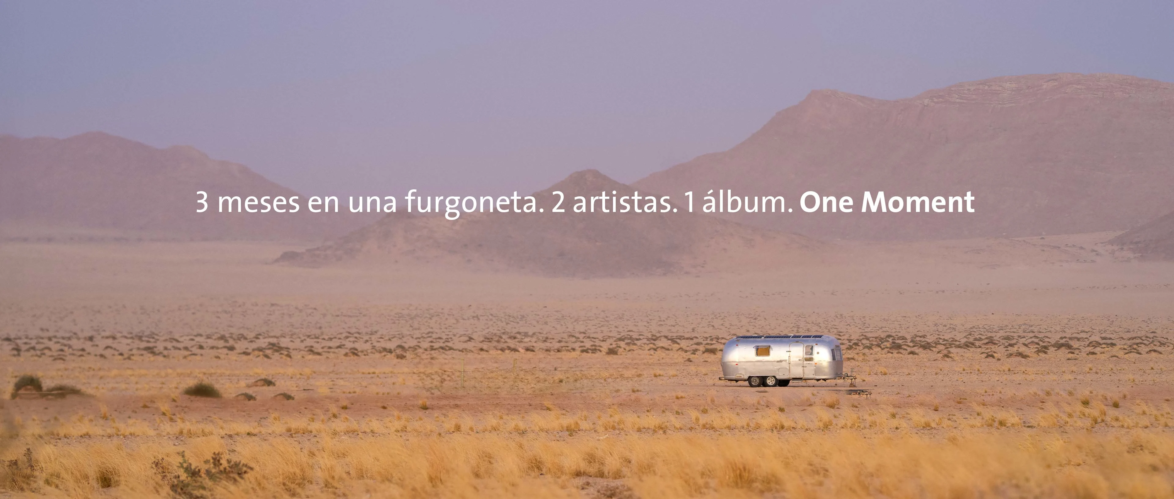 Una amplia toma panorámica del desierto de Namibia bajo una luz violeta difusa. En mitad de la llanura arenosa se encuentra una caravana Airstream plateada frente a una suave cadena montañosa. En el centro de la imagen aparece el texto: "3 meses en una furgoneta. 2 artistas. 1 álbum. One Moment".