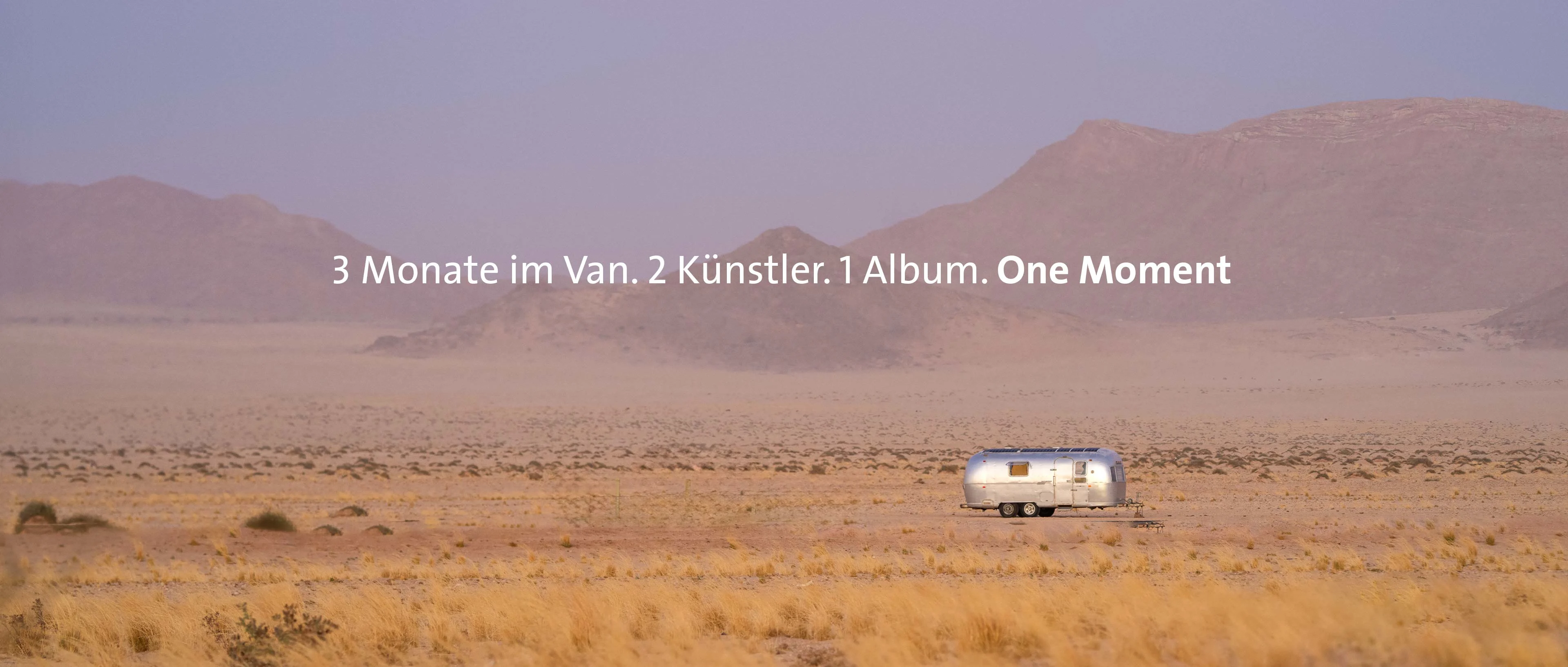 Eine weite Panoramaaufnahme der namibischen Wüste bei diffusem, violettem Licht. In der Mitte der sandigen Ebene steht ein silberner Airstream-Wohnwagen vor einer sanften Bergkette. Im Zentrum des Bildes steht der Text: „3 Monate in einem Van. 2 Künstler. 1 Album. One Moment“.