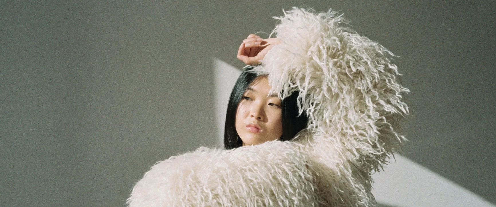 A woman with black hair and a white, long-haired fur cloak raises one arm against a light-colored background.