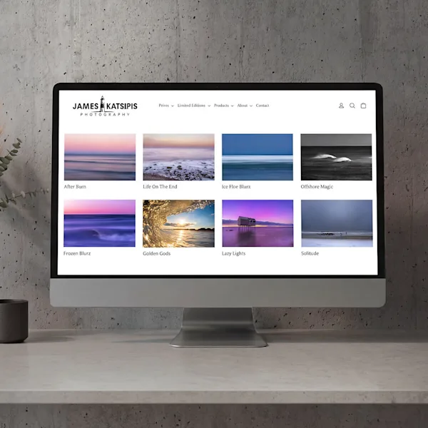 Ein silberner Desktop-Monitor steht auf einem hellen Tisch vor einer Sichtbetonwand. Auf dem Bildschirm ist die Webseite „James Katsipis Photography“ zu sehen, die eine Galerie mit acht verschiedenen Landschafts- und Meeresaufnahmen in stimmungsvollen Farben zeigt. Links im Bild ist dezent ein Teil einer Pflanze in einem dunklen Topf erkennbar.