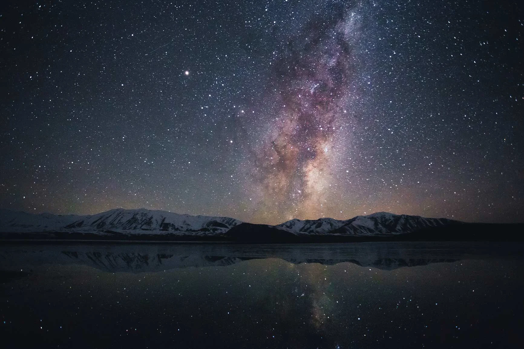 A starry sky with the Milky Way above snow-capped mountains, reflected in a calm lake.