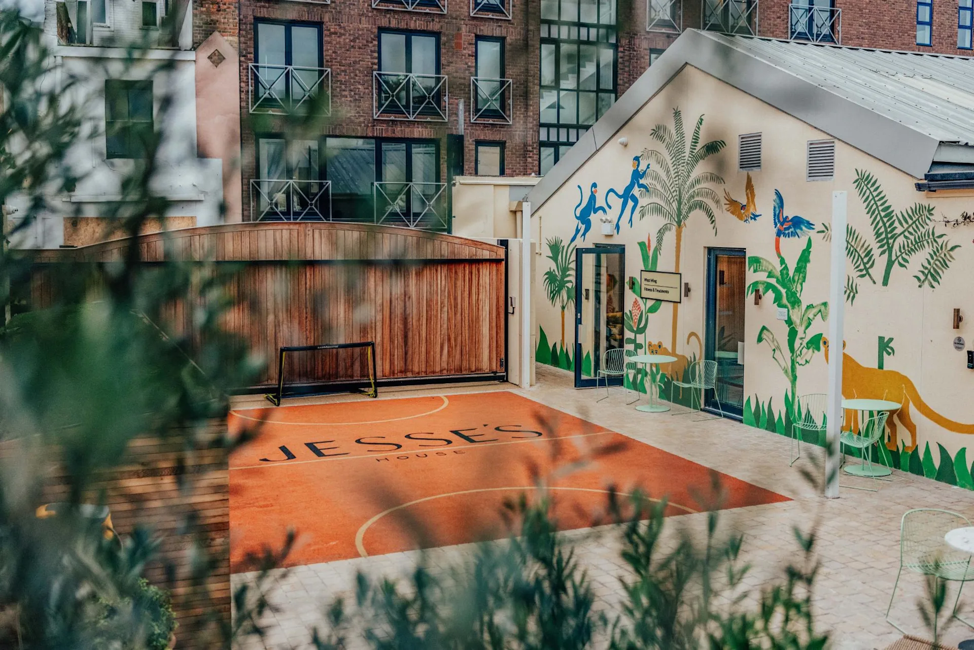 A small outdoor basketball court labeled Jesses House is surrounded by greenery. The adjacent building features colorful, playful animal murals. Apartment buildings are visible in the background.