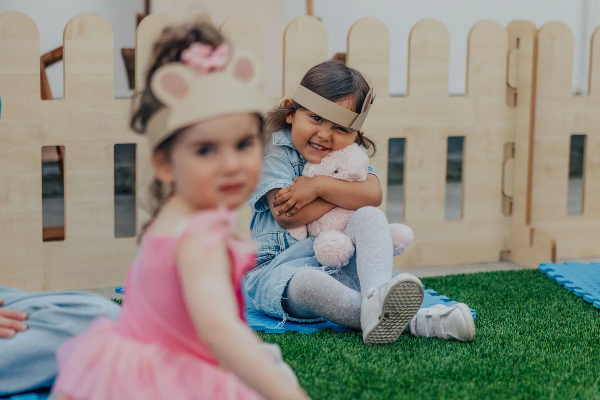 A young girl in a blue dress sits on artificial grass, hugging a stuffed animal and smiling. Another child in a pink outfit and animal ear headband is blurred in the foreground. A wooden fence is in the background.