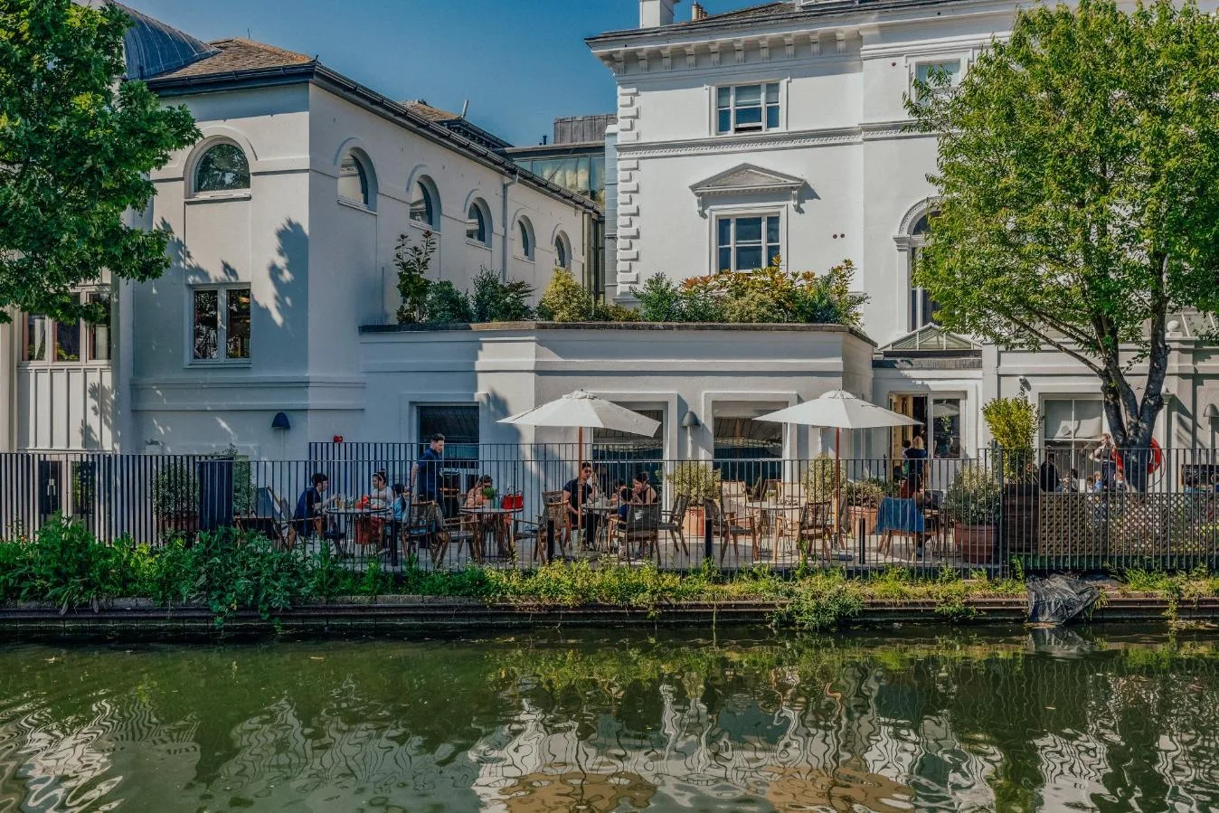 Outdoor café with people sitting under white umbrellas beside a canal, next to a white building with greenery and trees, on a sunny day.