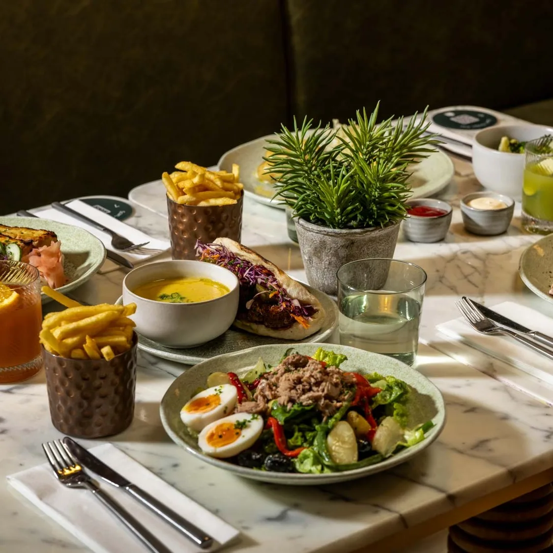 A marble table set with plates of salad, soup, bread, French fries, a wrap, drinks, and small potted plants in the center, ready for a meal in a restaurant setting.