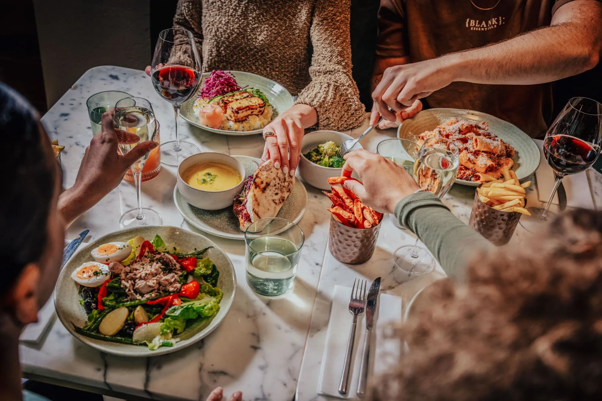 Four people dine together at a marble table set with salads, soup, main dishes, fries, and glasses of wine, reaching for food and sharing a meal in a cozy, well-lit setting.