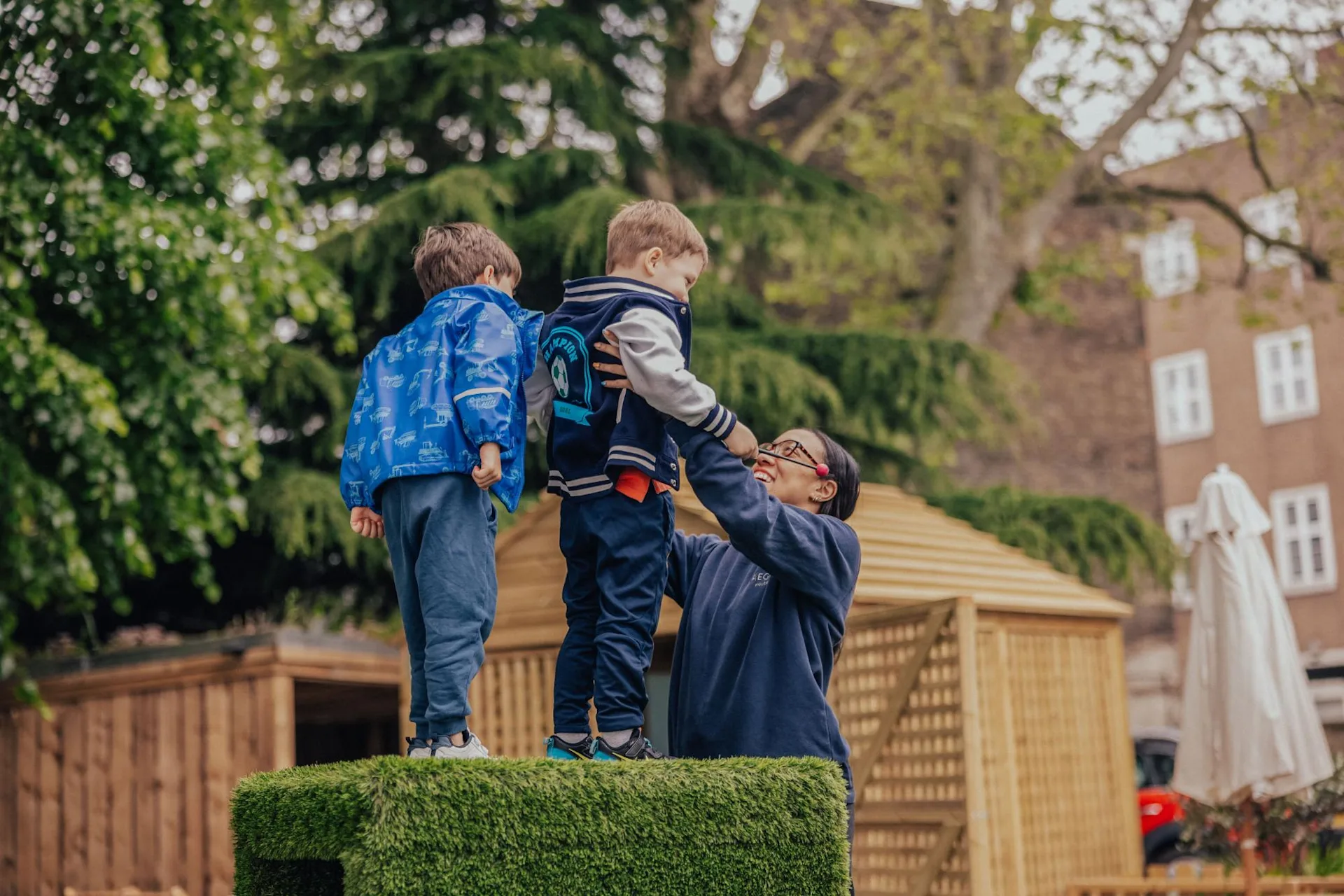 An adult helps lift a smiling boy off a grassy platform while another child stands nearby. They are outdoors, surrounded by trees and wooden structures.