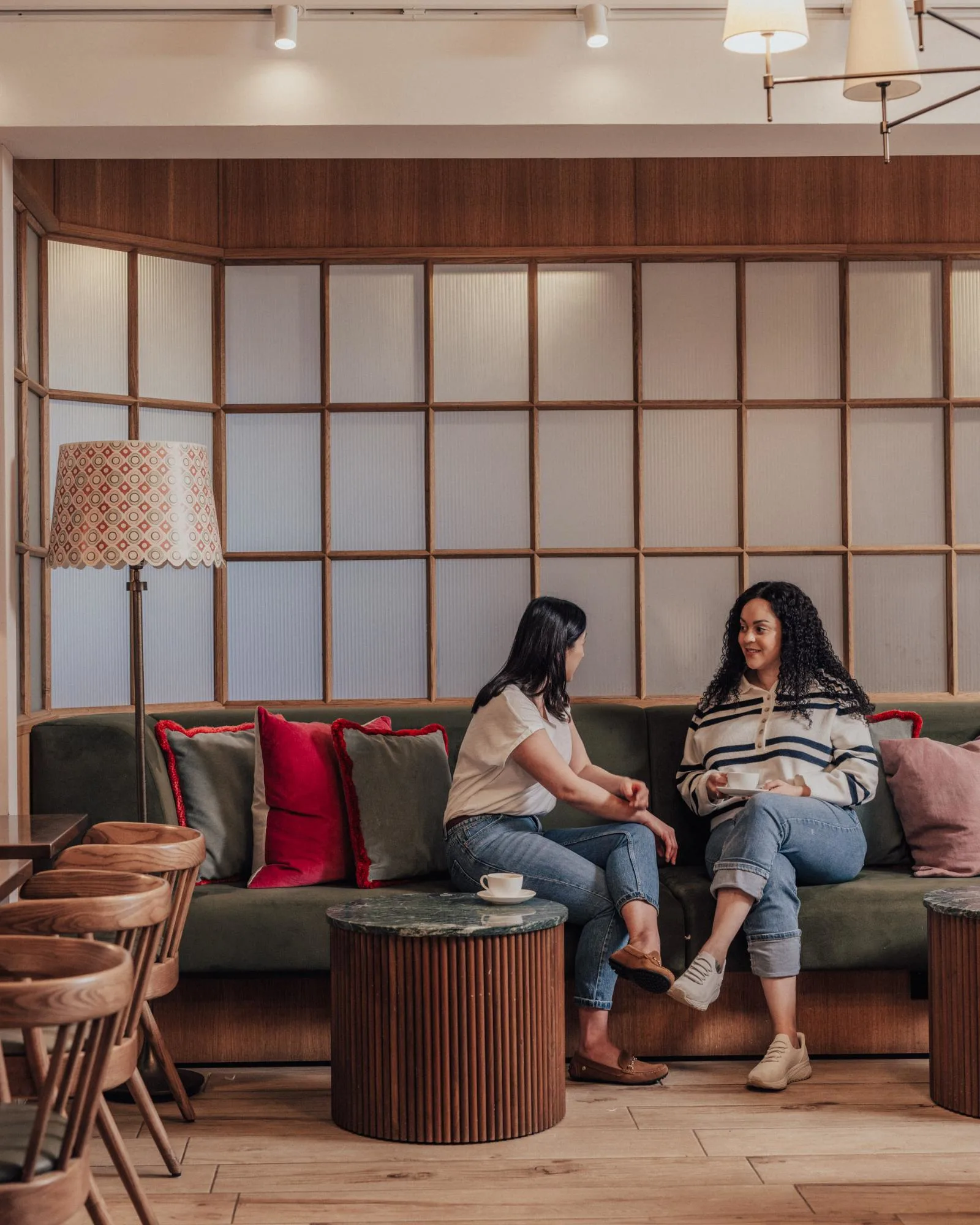 Two women are sitting on a sofa in a cozy café, engaged in conversation. They are surrounded by decorative pillows, wooden tables, and chairs, with a cup of coffee on the table in front of them.