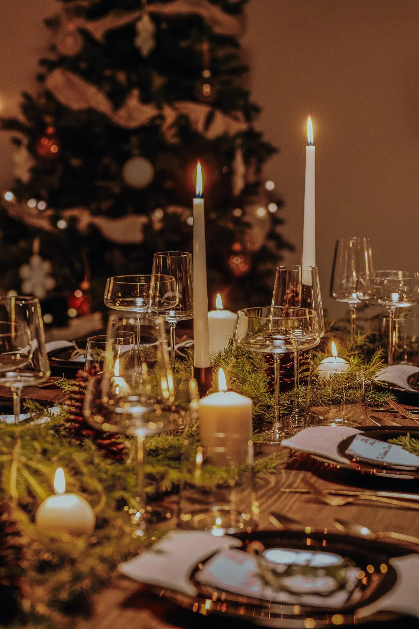 A warmly lit dining table set for a festive meal with candles, glassware, pinecones, and greenery. A decorated Christmas tree with ornaments and lights is visible in the background.