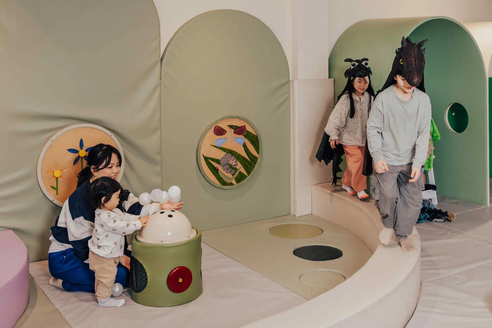 Children play in a soft, indoor play area; one child presses a bubble-making toy with an adult, while two kids in animal costumes walk nearby, all surrounded by pastel-colored padded walls and playful decorations.