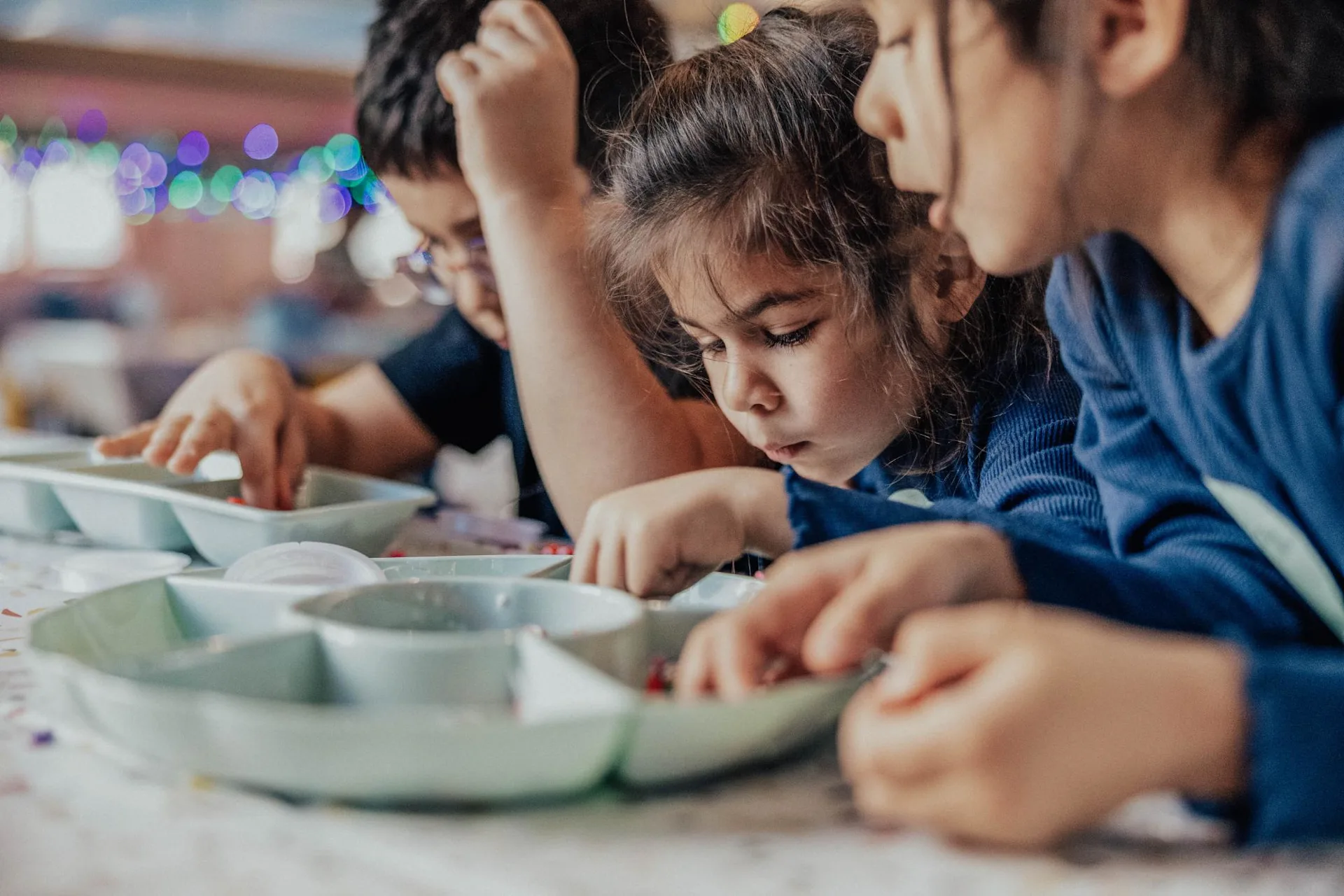 Three young children are focused on sorting small objects or beads in divided trays on a table, likely participating in an arts and crafts activity. Colorful lights blur in the background.
