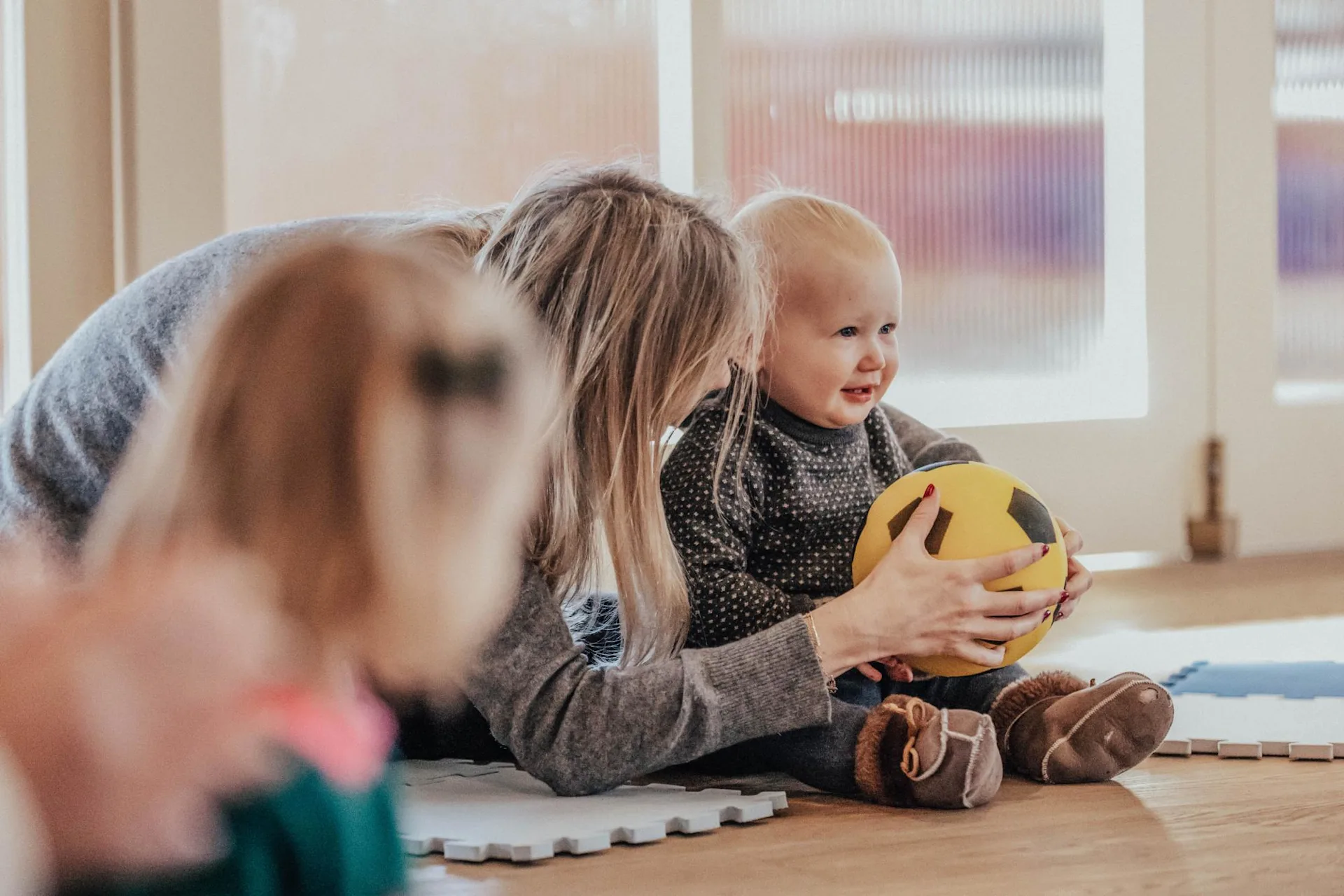 A woman helps a smiling toddler hold a yellow and black ball while sitting on a play mat indoors. Another young child is blurred in the foreground.