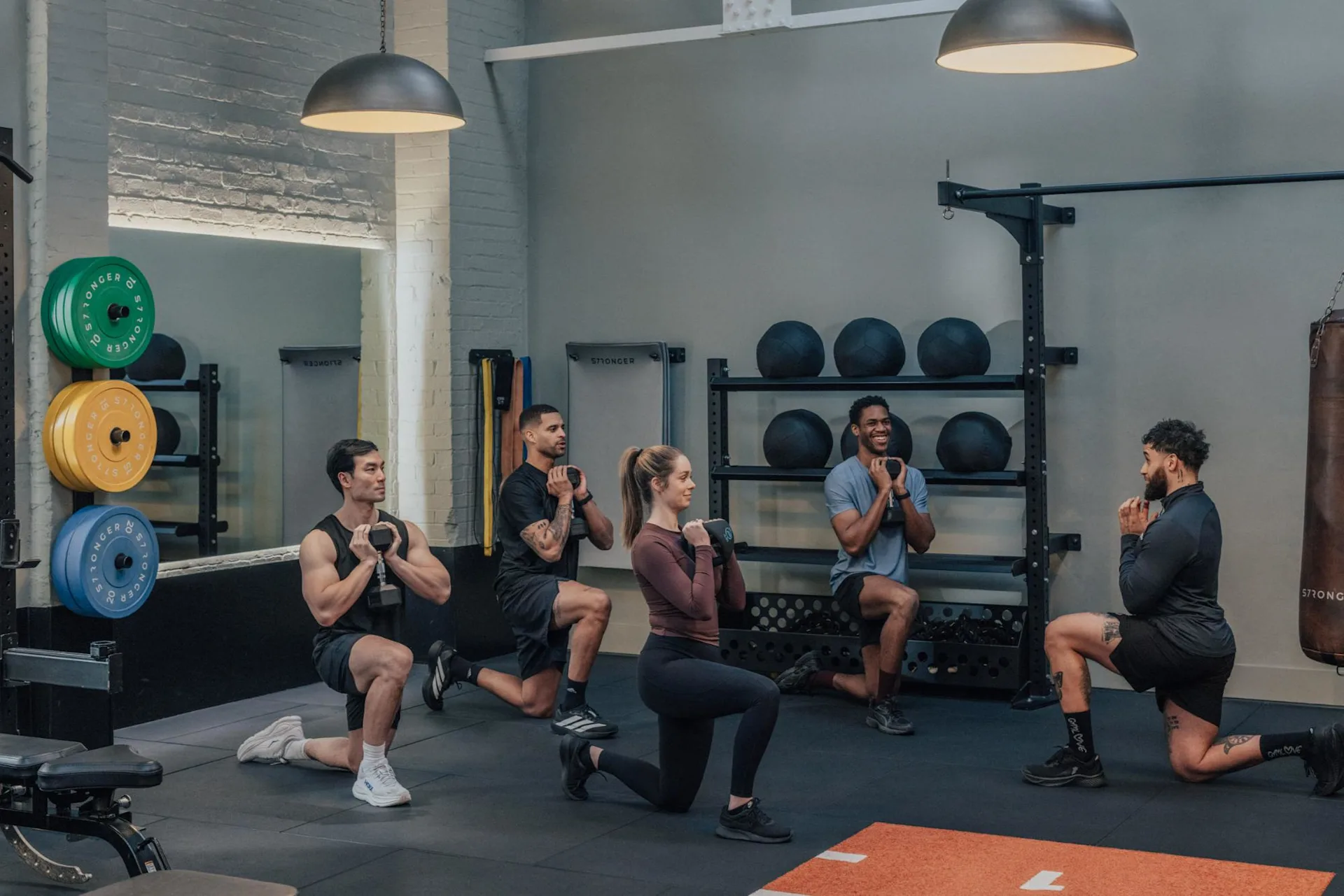 Five people in athletic wear perform weighted lunges together in a gym, following an instructor. Weight plates, medicine balls, and gym equipment are visible in the background.