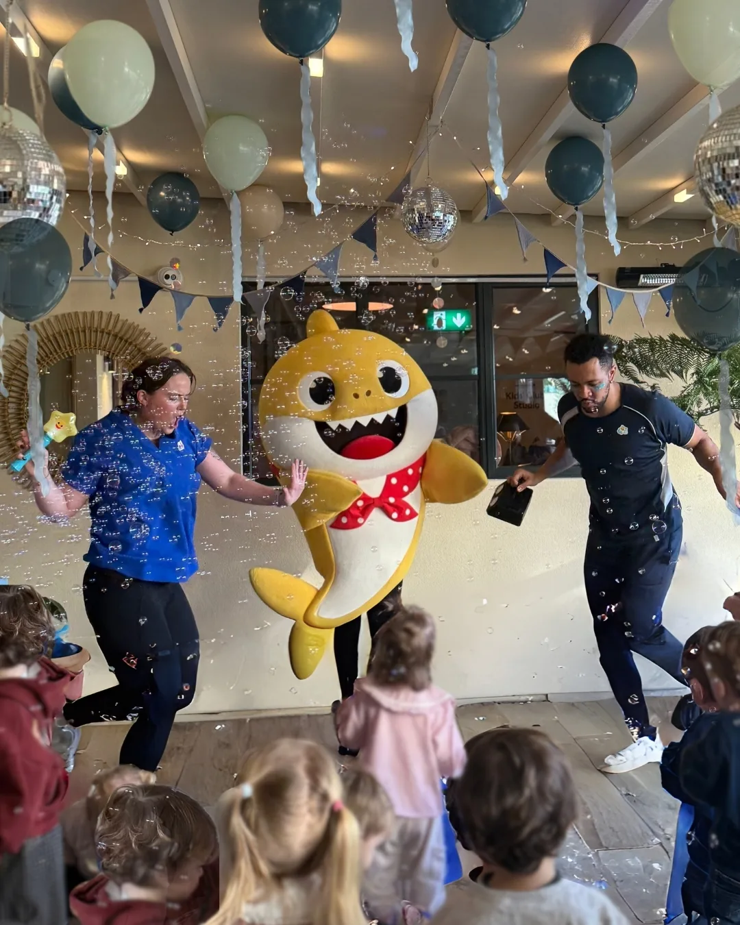 Three adults, including one in a yellow shark costume with a red bow tie, dance among bubbles and children at a party decorated with balloons, bunting, and disco balls. Children watch and join in the fun.