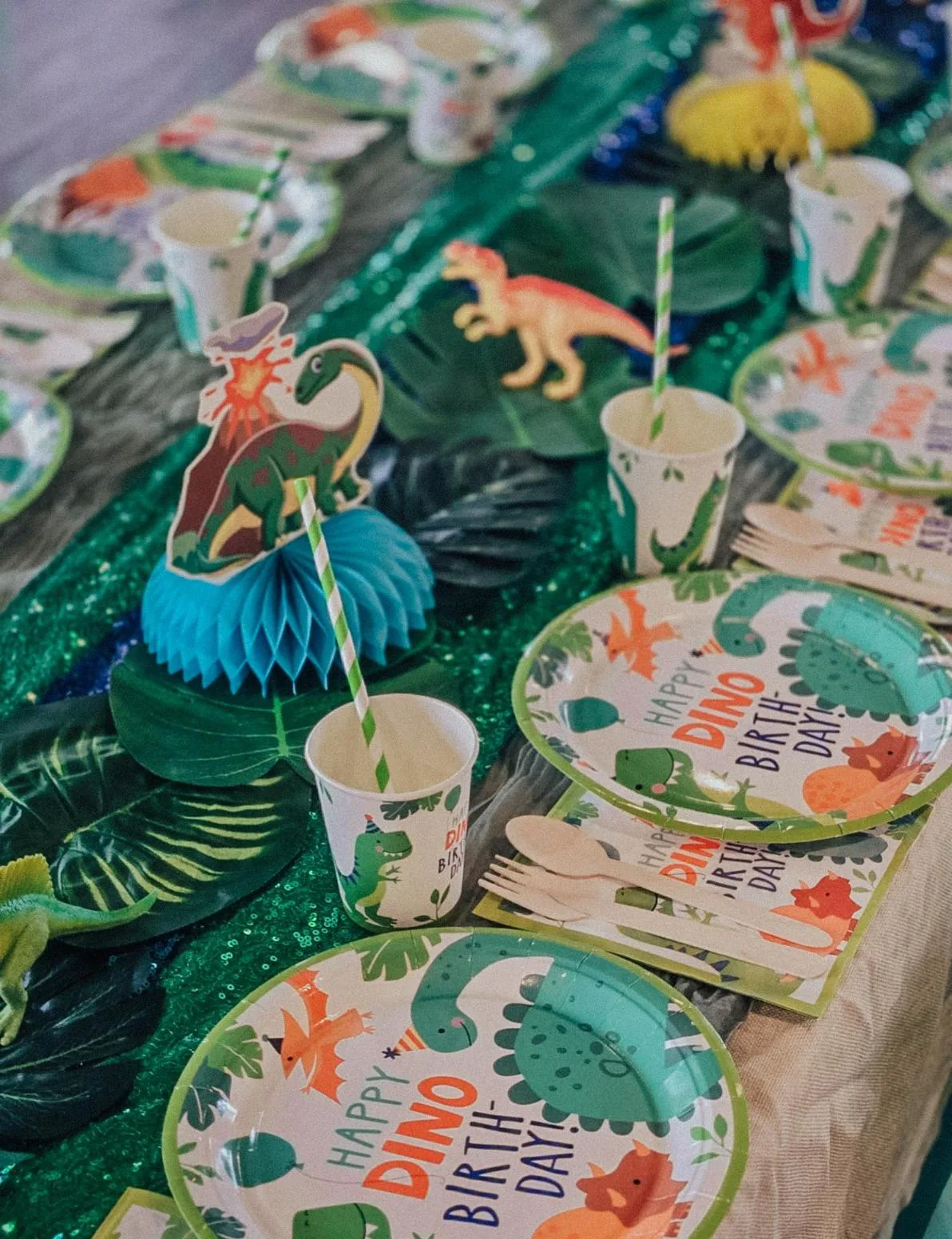 A festive table set for a dinosaur-themed birthday party, featuring plates, cups, napkins, and straws decorated with cartoon dinosaurs, along with dinosaur figurines and leafy decorations as the centerpiece.