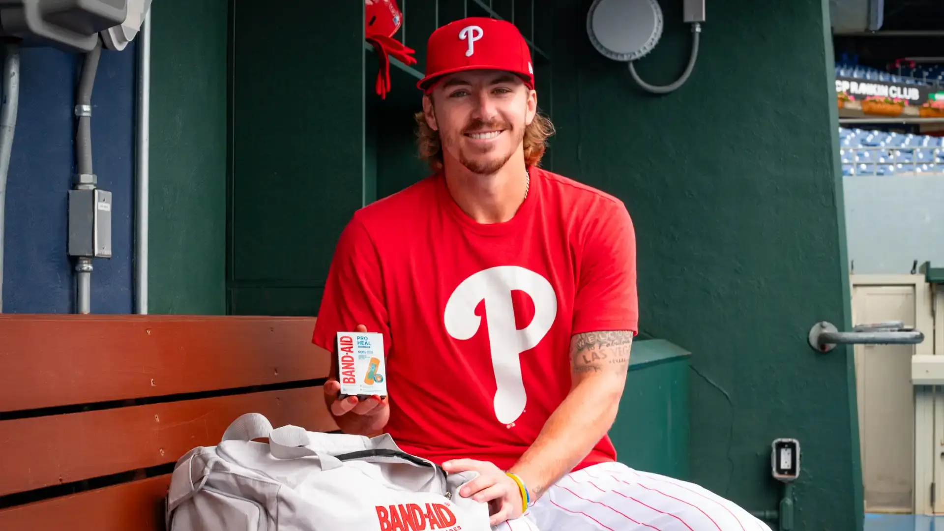 Bryson Stott wearing a red Philadelphia Phillies shirt and cap stands in front of a large screen at Citizens Bank Park, holding a package of Band-Aid Pro Heal. The baseball field is visible in the background.