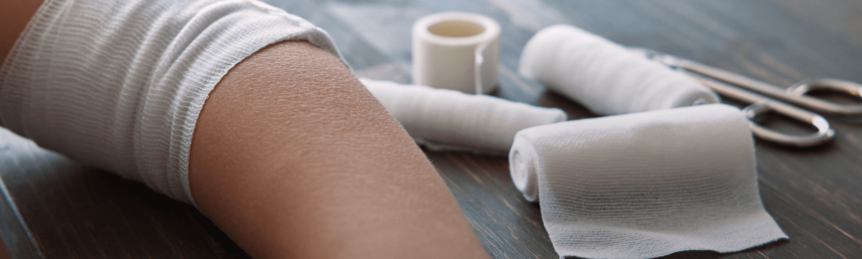 Person’s bandaged arm resting on table next to gauze wrap and medical tape. 