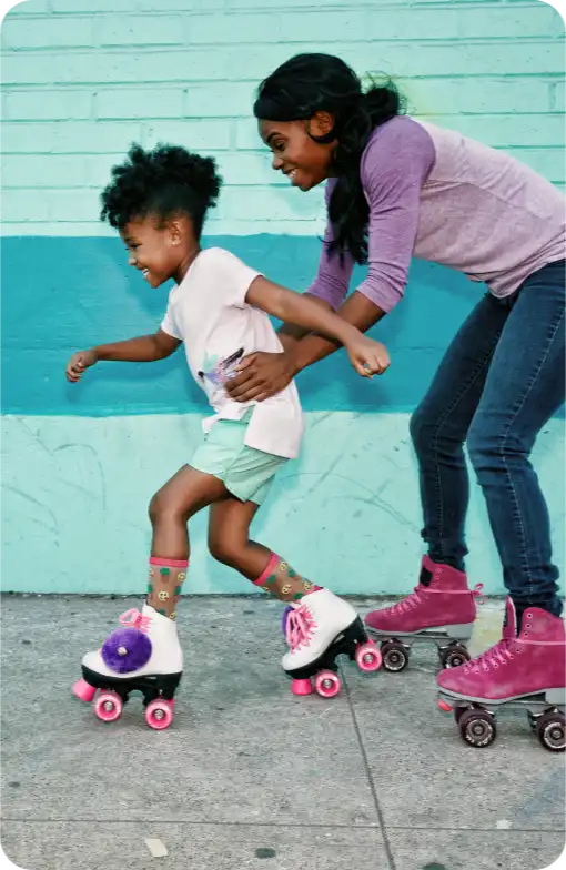 A woman and a child roller skating together on a sidewalk, both smiling and having fun.