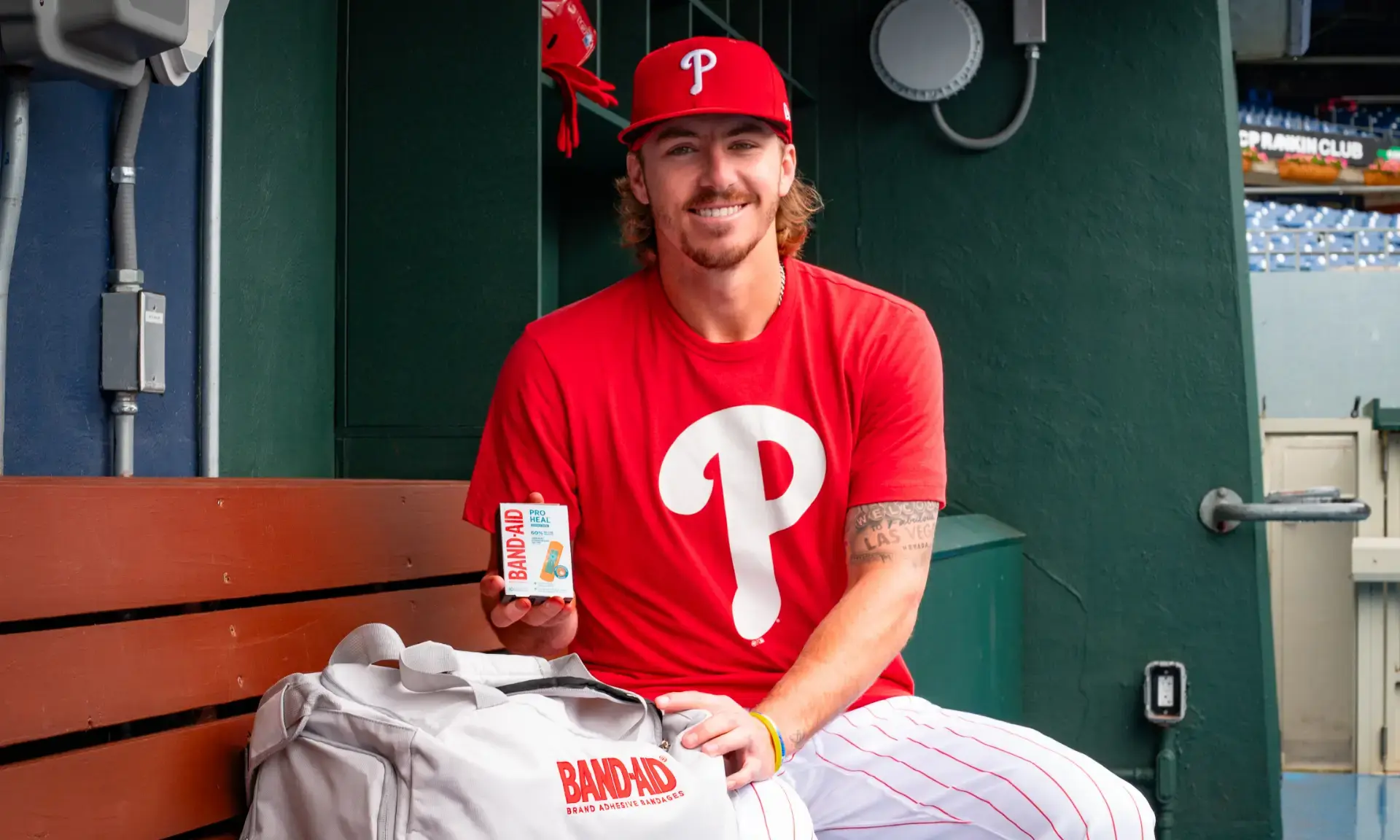 Bryson Stott wearing a red Philadelphia Phillies shirt and cap stands in front of a large screen at Citizens Bank Park, holding a package of Band-Aid Pro Heal. The baseball field is visible in the background.