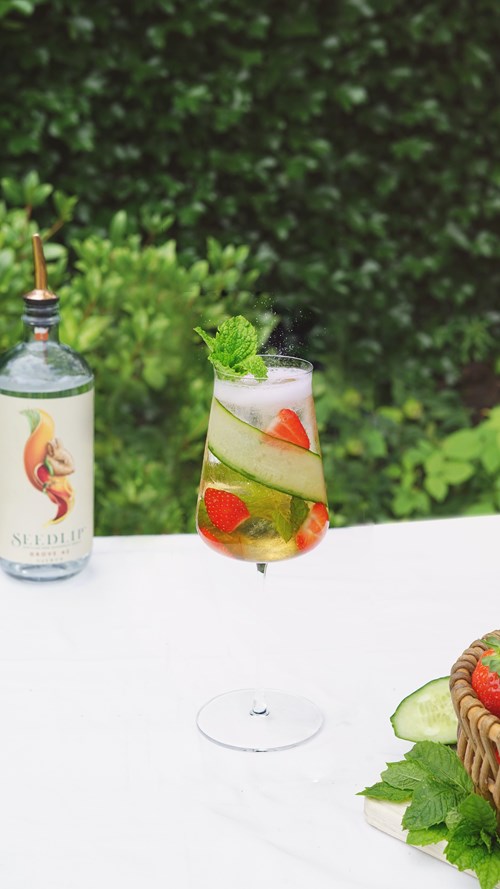 Refreshing Grove Fruit Cup with Seedlip Grove 42 garnished with cucumber slices strawberries and mint leaves on a white table with greenery background