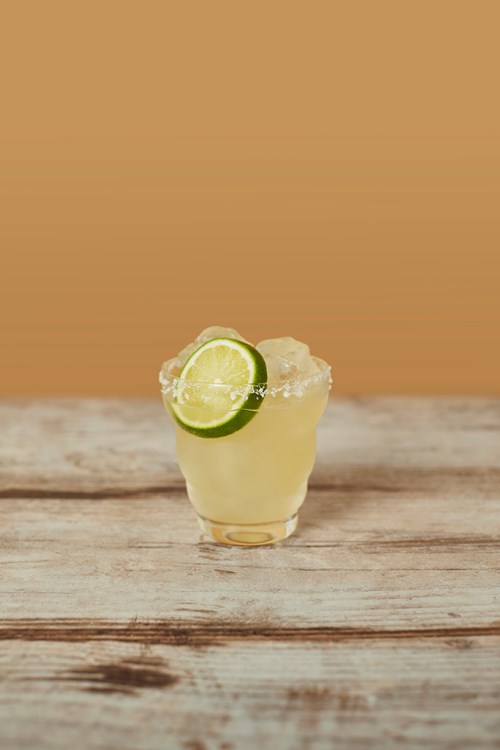 Seedlip Grove 42 Grove Margarita cocktail with ice cubes, lime wheel garnish, and salted rim, on a rustic wooden surface against an orange background.