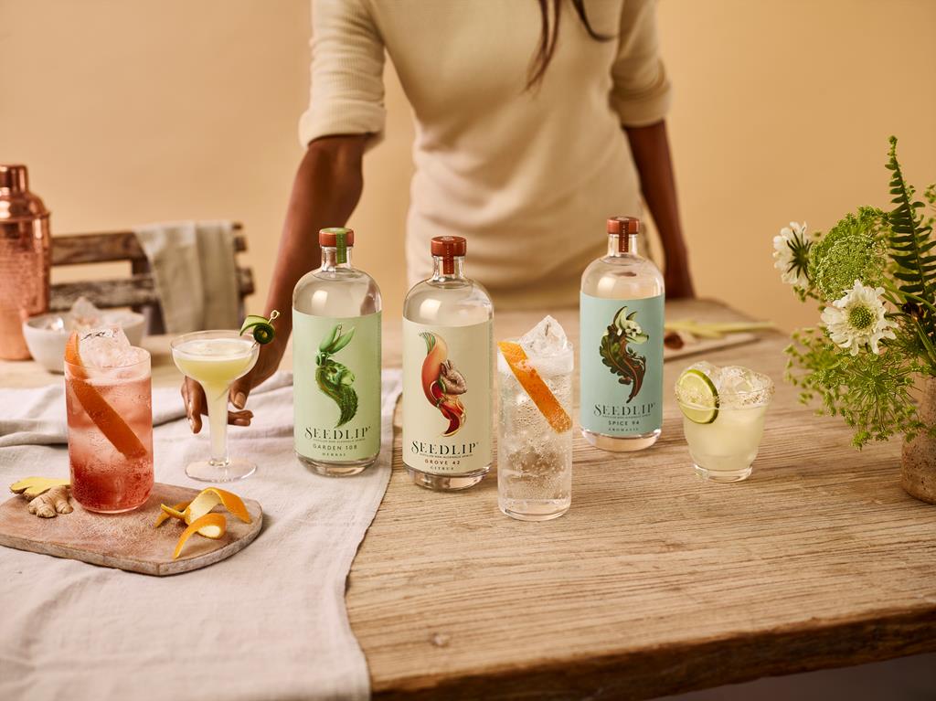 Person behind a wooden table with three Seedlip non-alcoholic spirits, cocktails, garnishes, a copper shaker, and flowers.