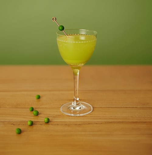 Peatini cocktali in a small cocktail glass on a wood counter with some peas scattered along side the base of the glass