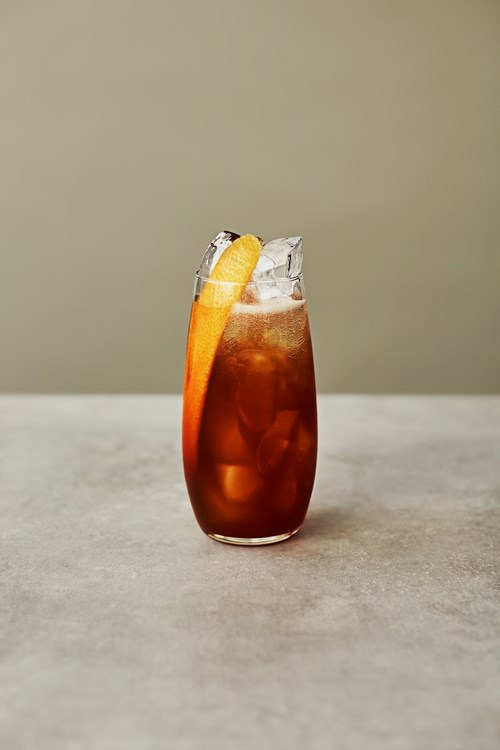 Tall glass filled with dark brown cold brew Seedlip Spice 94 cocktail, garnished with a long orange peel and ice cubes, placed on a light gray surface