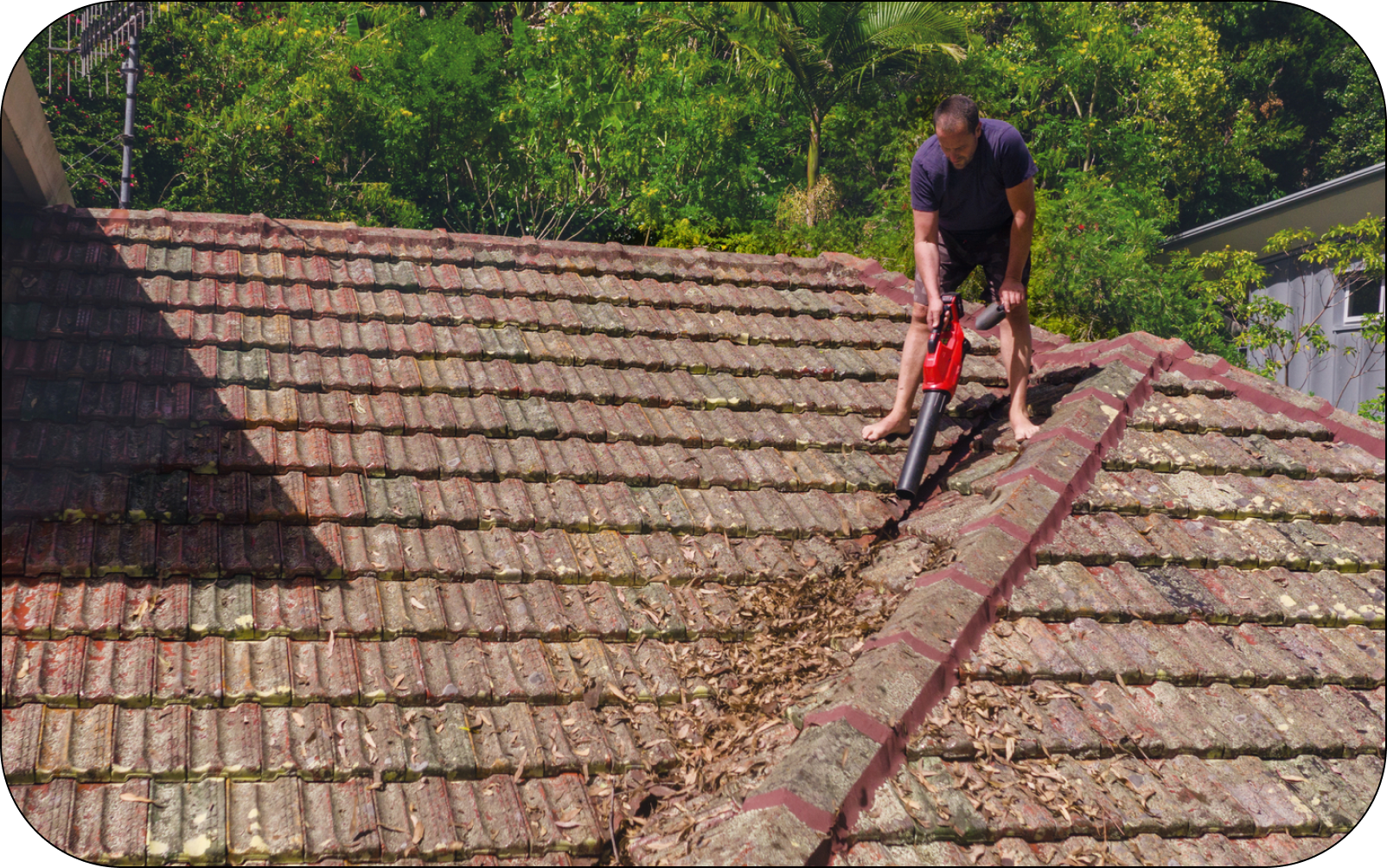 Image of a property owner on the roof of a house and using a leaf blower to clear the gutters 