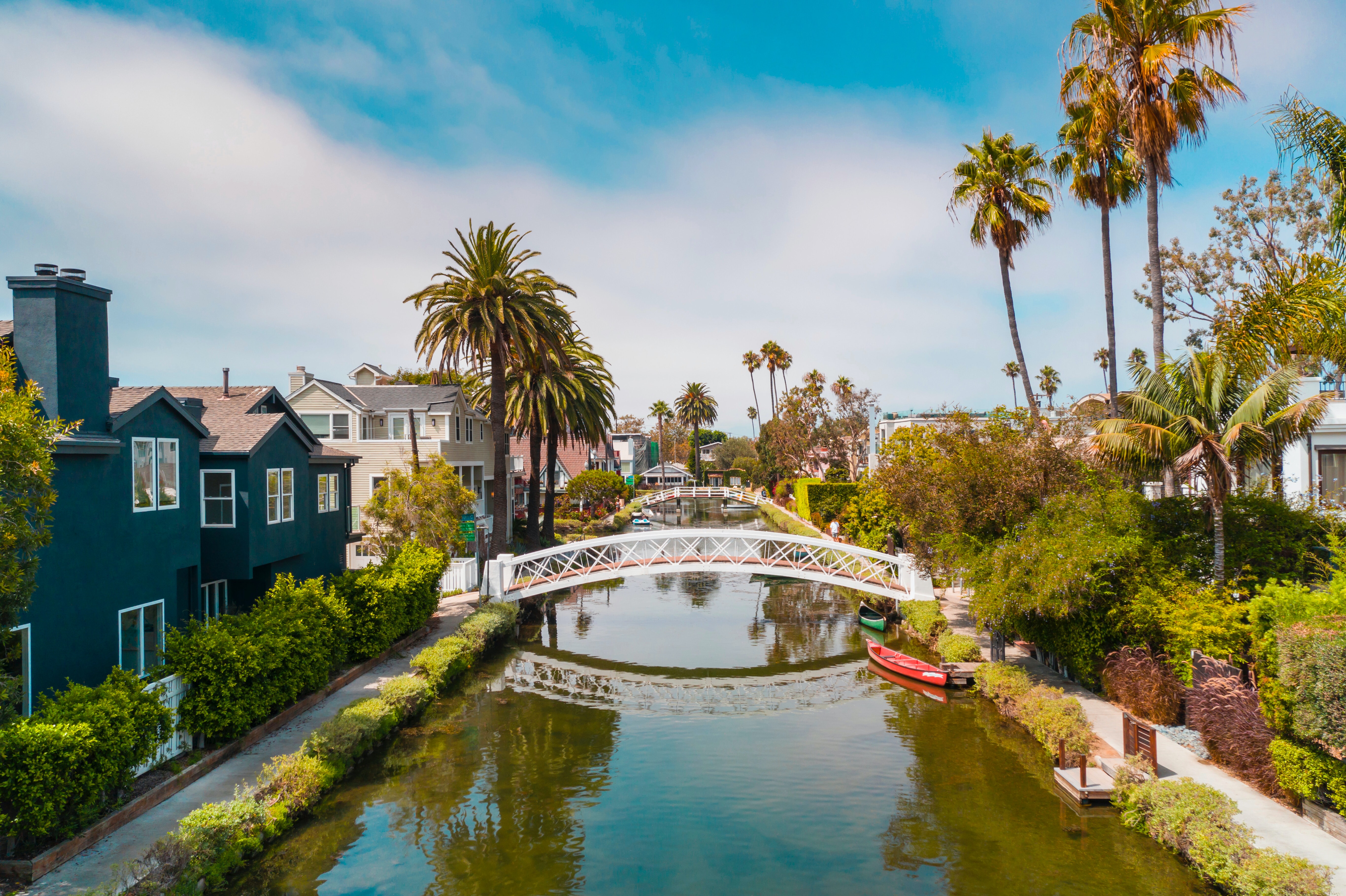 A photo of one of the waterways in Venice california with a white bridge going over the water from left to right. Houses on either side of the river and palm trees in the background.