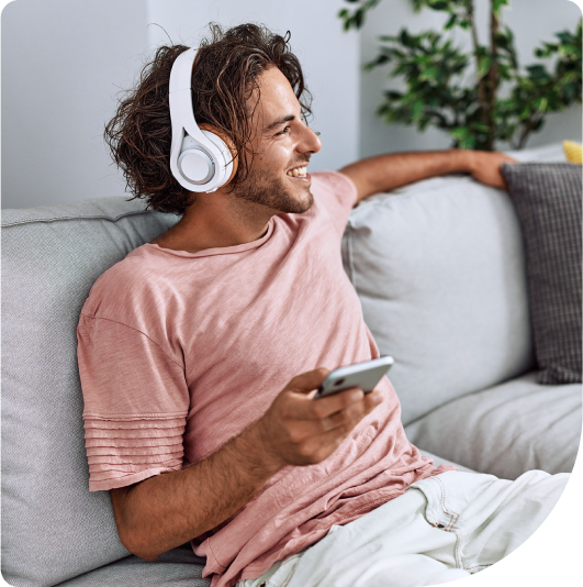 An image of a smiling man sitting on a grey couch and listening to music on white headphones