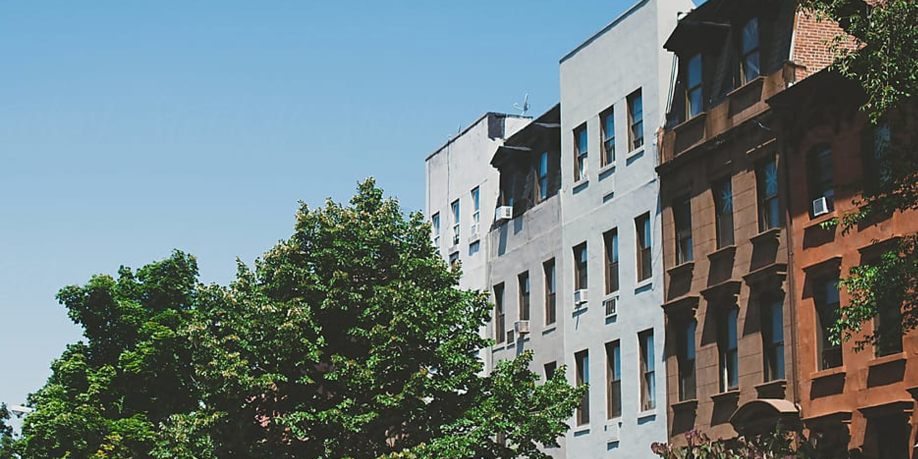 A row of apartment buildings and trees against a blue sky. 