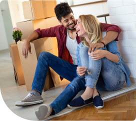 An image of a couple sitting on the floor with moving boxes around them, smiling at each other