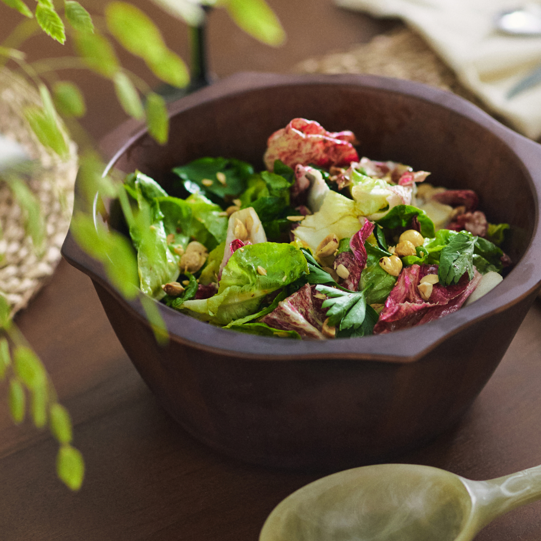 Fresh mixed greens salad with nuts in a dark brown bowl, with green plant leaves visible in the background.