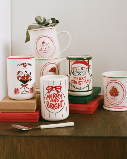 Collection of white ceramic holiday mugs with red festive designs including Santa, wine glass, bow, and Christmas tree on stacked books