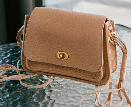 Tan leather crossbody purse with gold turnlock clasp and adjustable strap on dark green marble surface